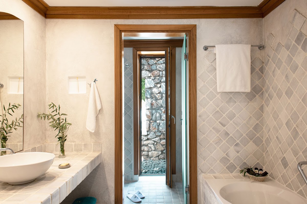 Spa-style bathroom with round vessel sink, diamond-pattern tile walls, wooden framed doorway opening to stone courtyard with greenery.