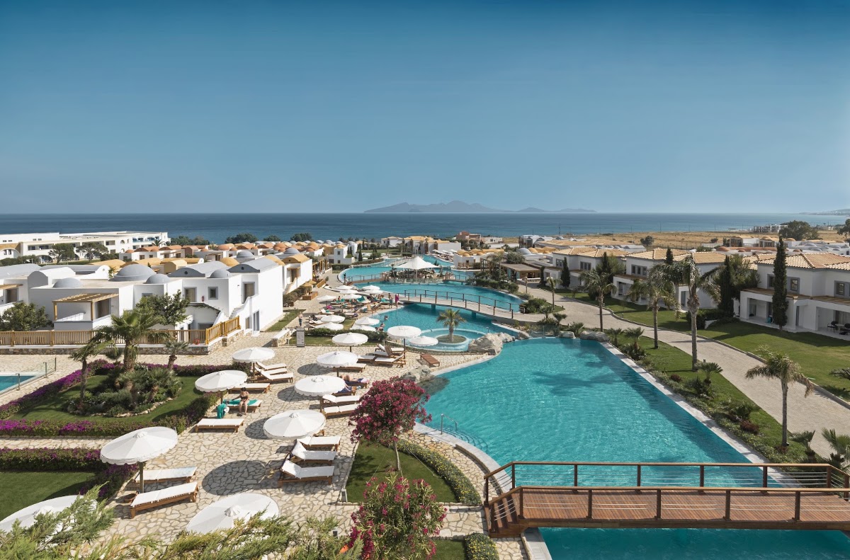 Aerial view of Mitsis Selection Blue Domes with turquoise pools, white villas, loungers with umbrellas, and the sea and mountains in the distance.