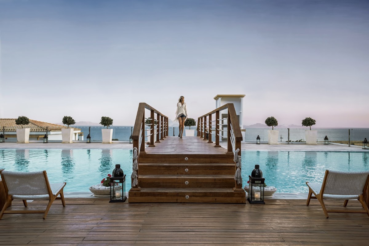 Woman walks down wooden boardwalk stairs flanked by infinity pools overlooking the Aegean Sea at Mitsis Selection Blue Domes.
