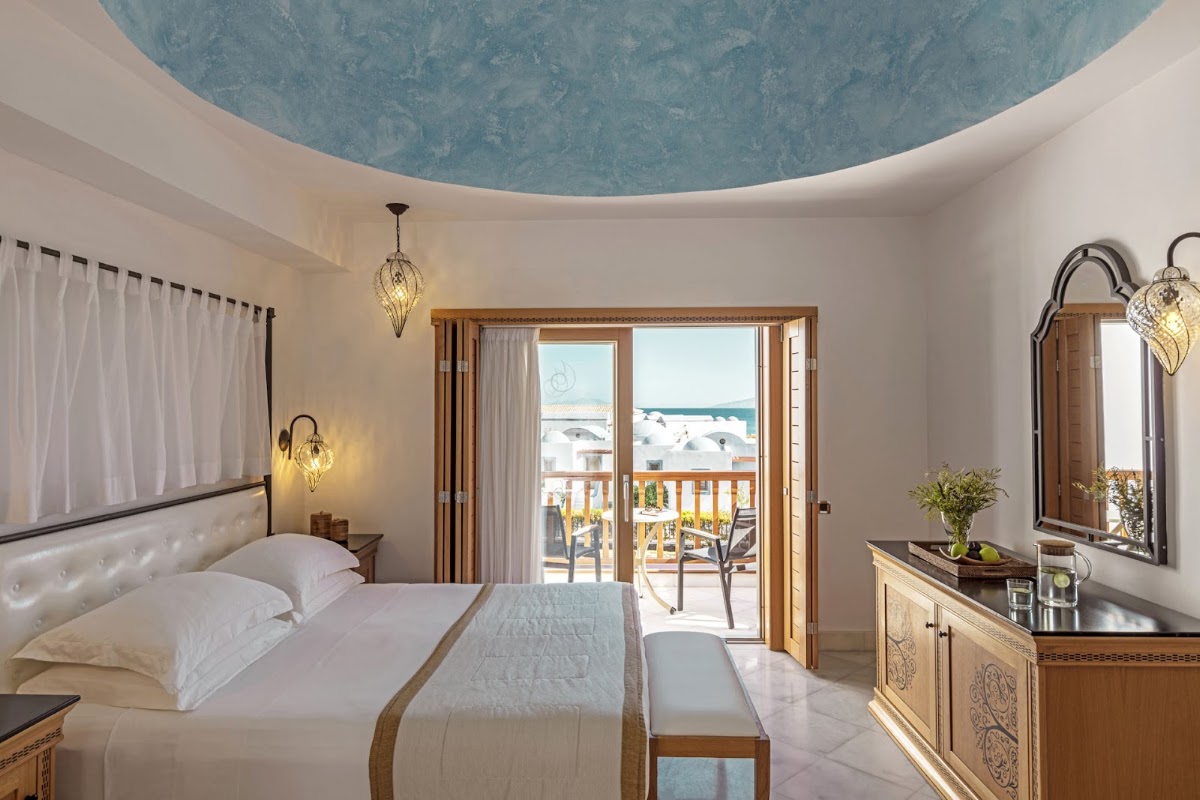 Resort bedroom with blue dome ceiling, white bedding, wooden furnishings, and sea view from balcony overlooking whitewashed buildings.