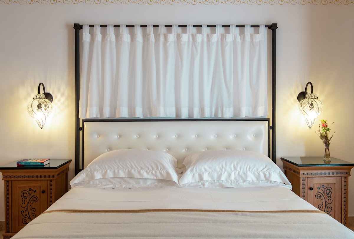Bedroom with black canopy bed frame, white tufted headboard, white pillows, and sheer curtains, flanked by carved wooden nightstands and decorative wall sconces.