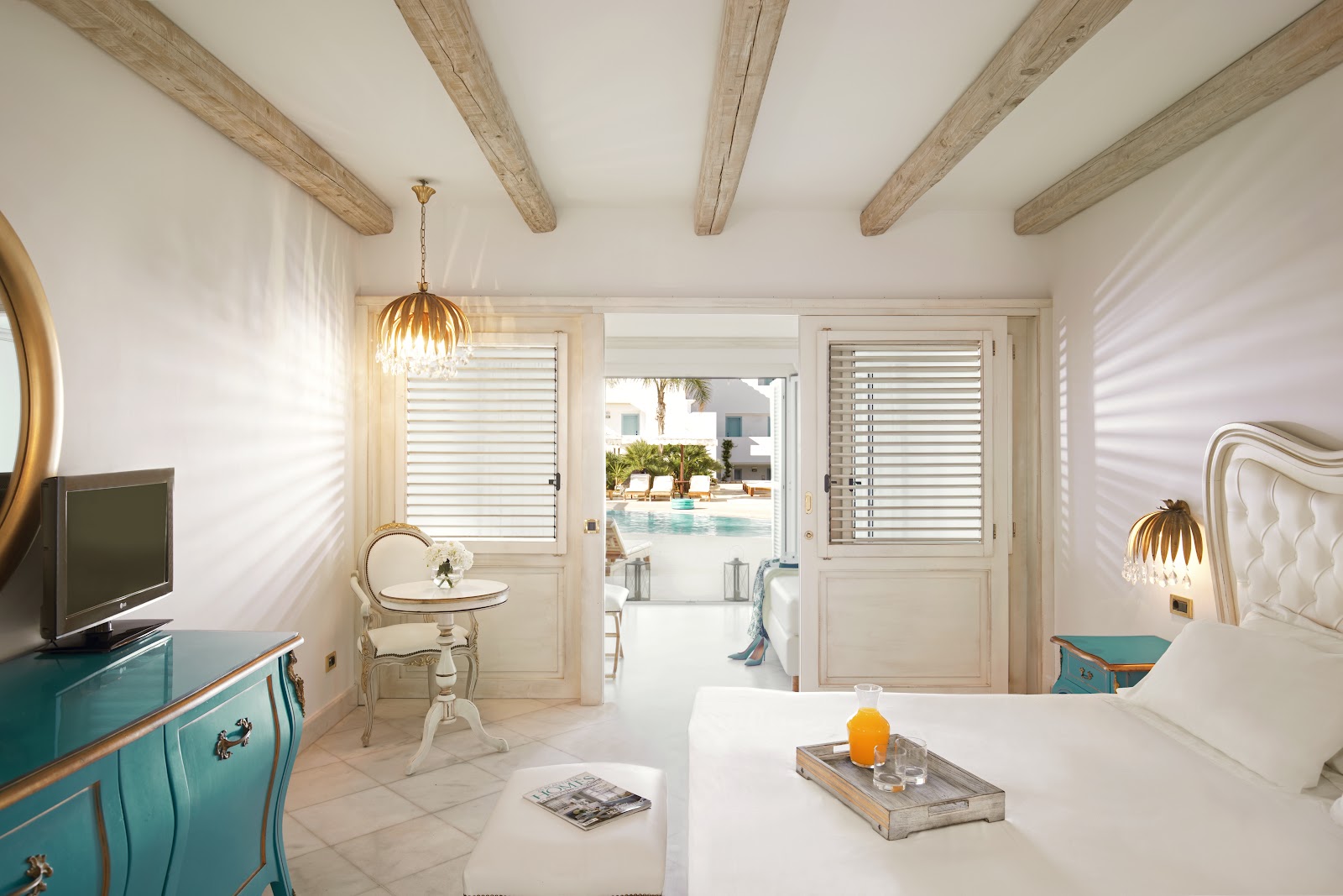 Bright bedroom with wooden beam ceiling, cream headboard, white bedding, turquoise dresser, and ocean view through open balcony doors.