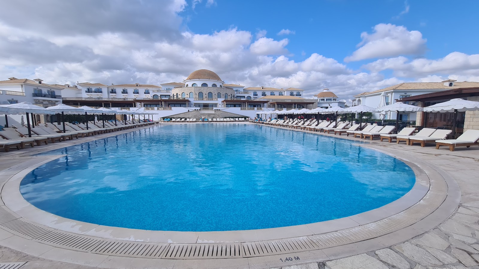 Large circular pool with loungers and umbrellas flanked by white resort buildings with domed roofs under partly cloudy sky.