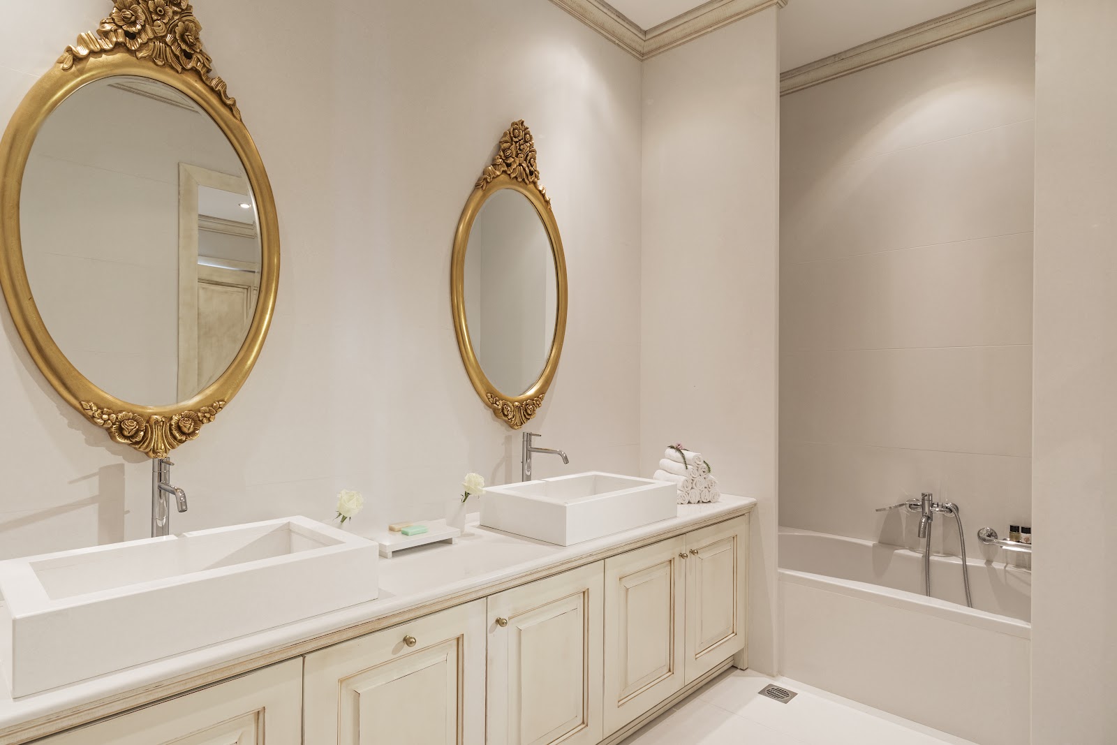 Bathroom with dual white sinks, cream cabinetry, and two ornate gold-framed oval mirrors on beige walls above the vanity.