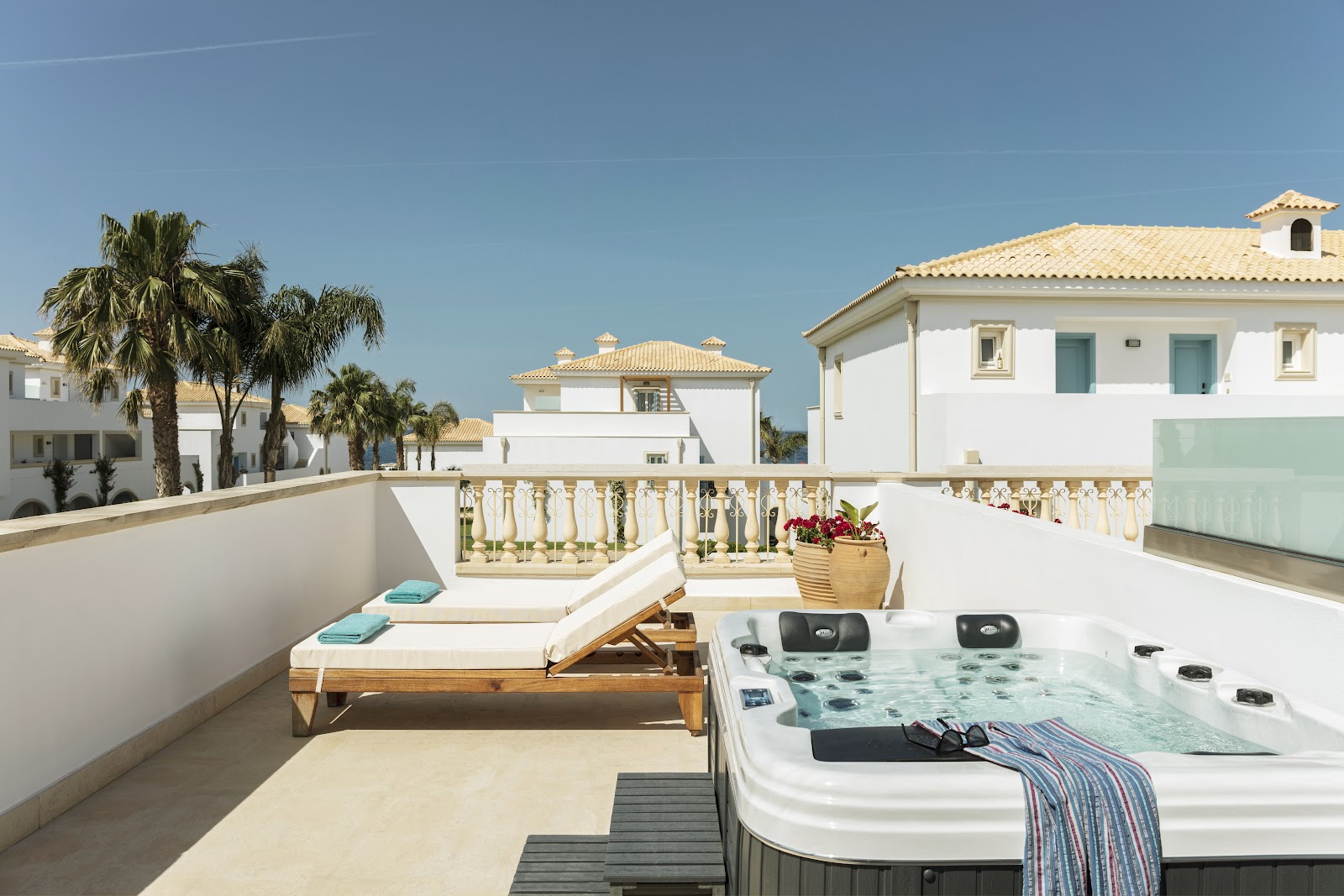 Stone building with arched windows overlooks a terrace where guests relax in lounge chairs and dining areas, with turquoise sea and sky beyond.