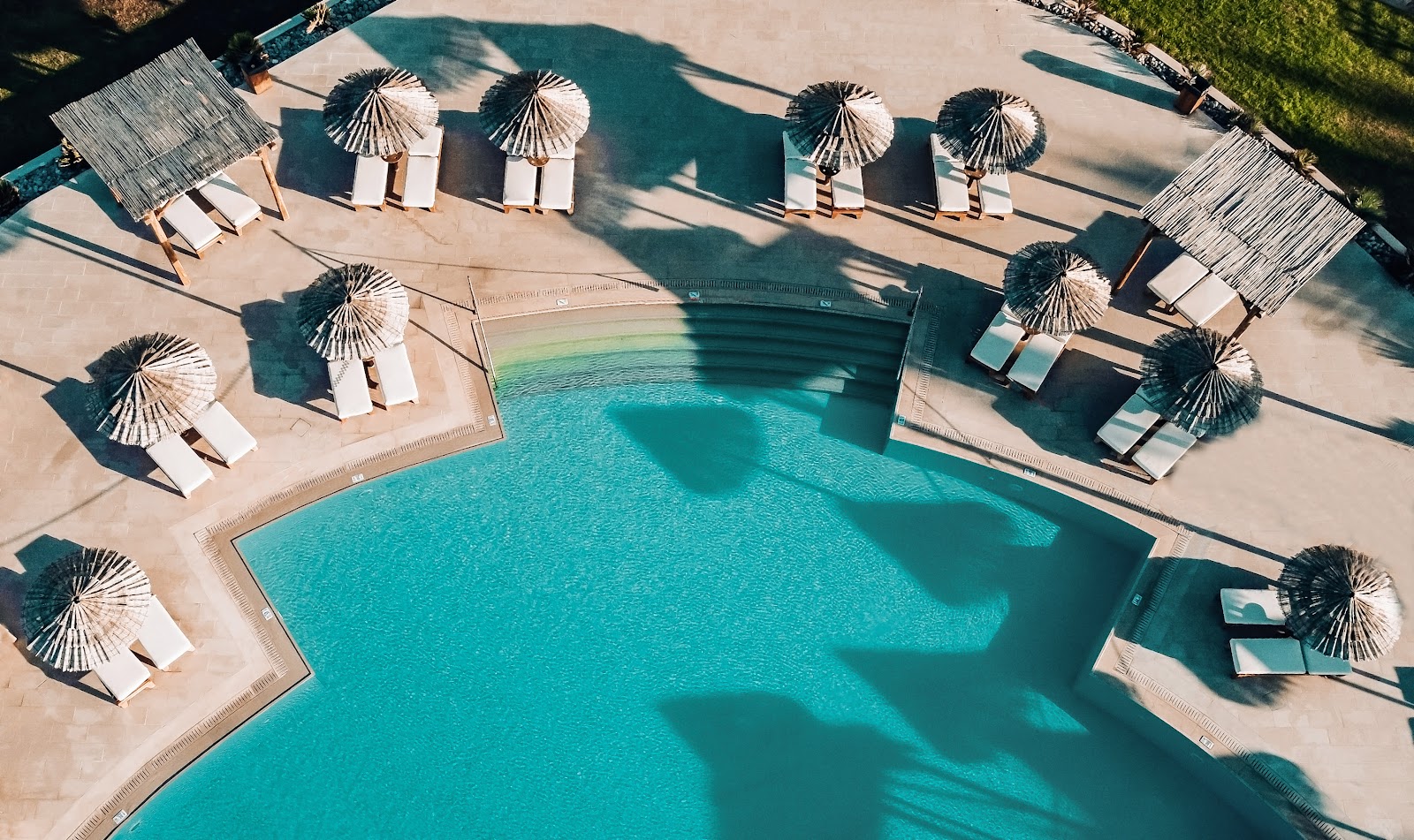 Overhead view of a resort pool with turquoise water surrounded by lounge chairs and thatched umbrellas casting shadows on the beige deck.