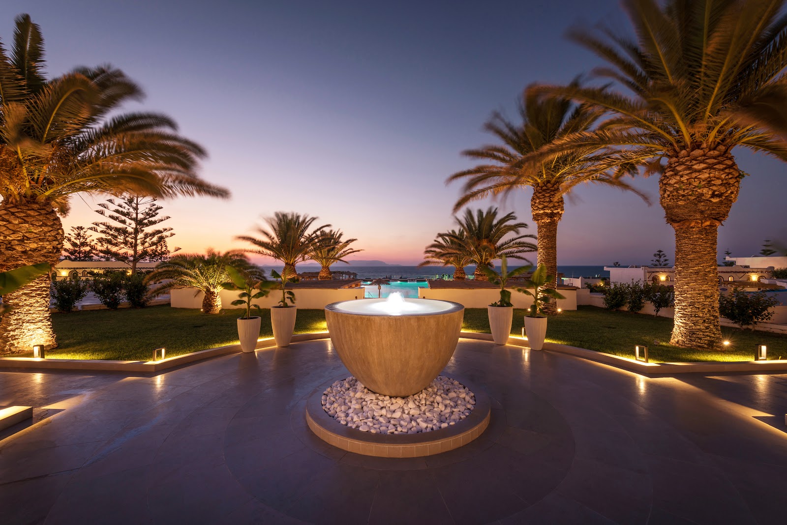 Illuminated wooden fire bowl surrounded by palm trees and manicured gardens at dusk, with pool visible beyond and sea in the distance.