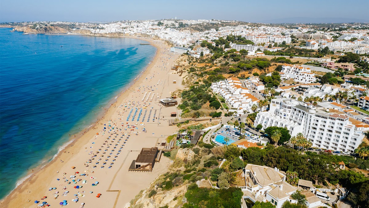 Aerial view of a curved sandy beach lined with blue umbrellas and sunbathers, turquoise water, and white buildings of the Apartamentos Monica Isabel Beach Club on the Portuguese coast.