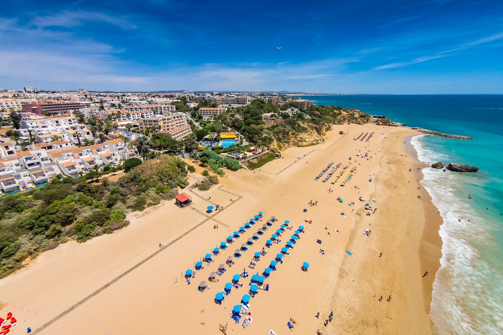 Aerial view of a sandy beach lined with blue umbrellas and sunbathers, bordered by turquoise ocean and a hillside resort with white buildings.