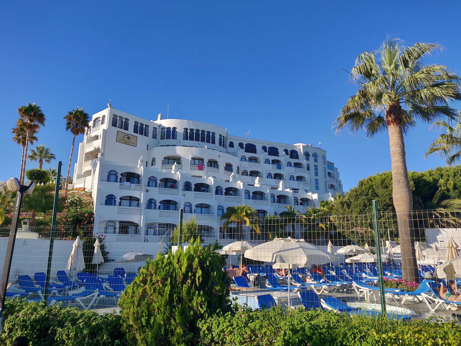 White Moorish-style resort building with blue balconies overlooking pool area with loungers and umbrellas, surrounded by palm trees.
