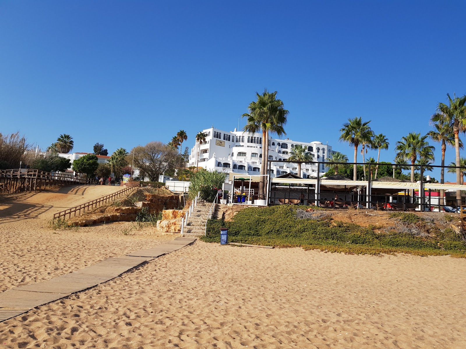 White multi-story resort building with palm trees and beach umbrellas under blue sky at Apartamentos Monica Isabel Beach Club.