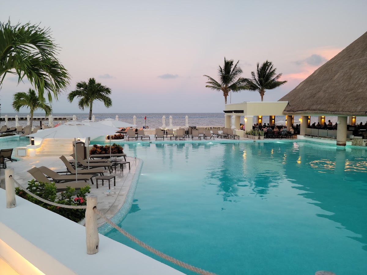 Large infinity pool at sunset with loungers, palm trees, thatched-roof bar, and ocean view beyond at Moon Palace Cancun.