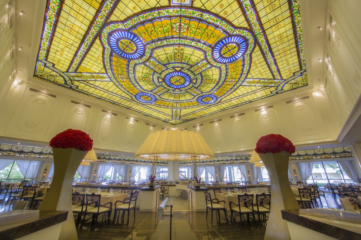 Dining room with ornate stained-glass ceiling featuring blue and yellow geometric patterns, red rose arrangements, and tables with black chairs.