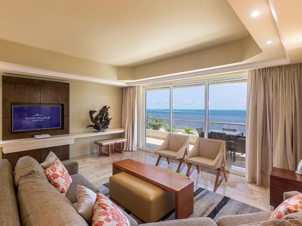 Moon Palace Cancun suite living room with ocean view through floor-to-ceiling windows, flat-screen TV, and coastal furniture.