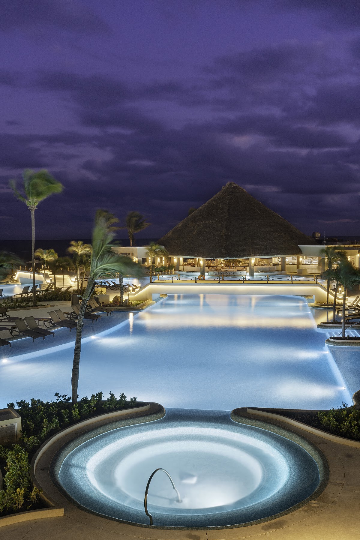 Illuminated pool complex at dusk with circular hot tub in foreground, pyramid-roofed main building, and palm trees under stormy sky.