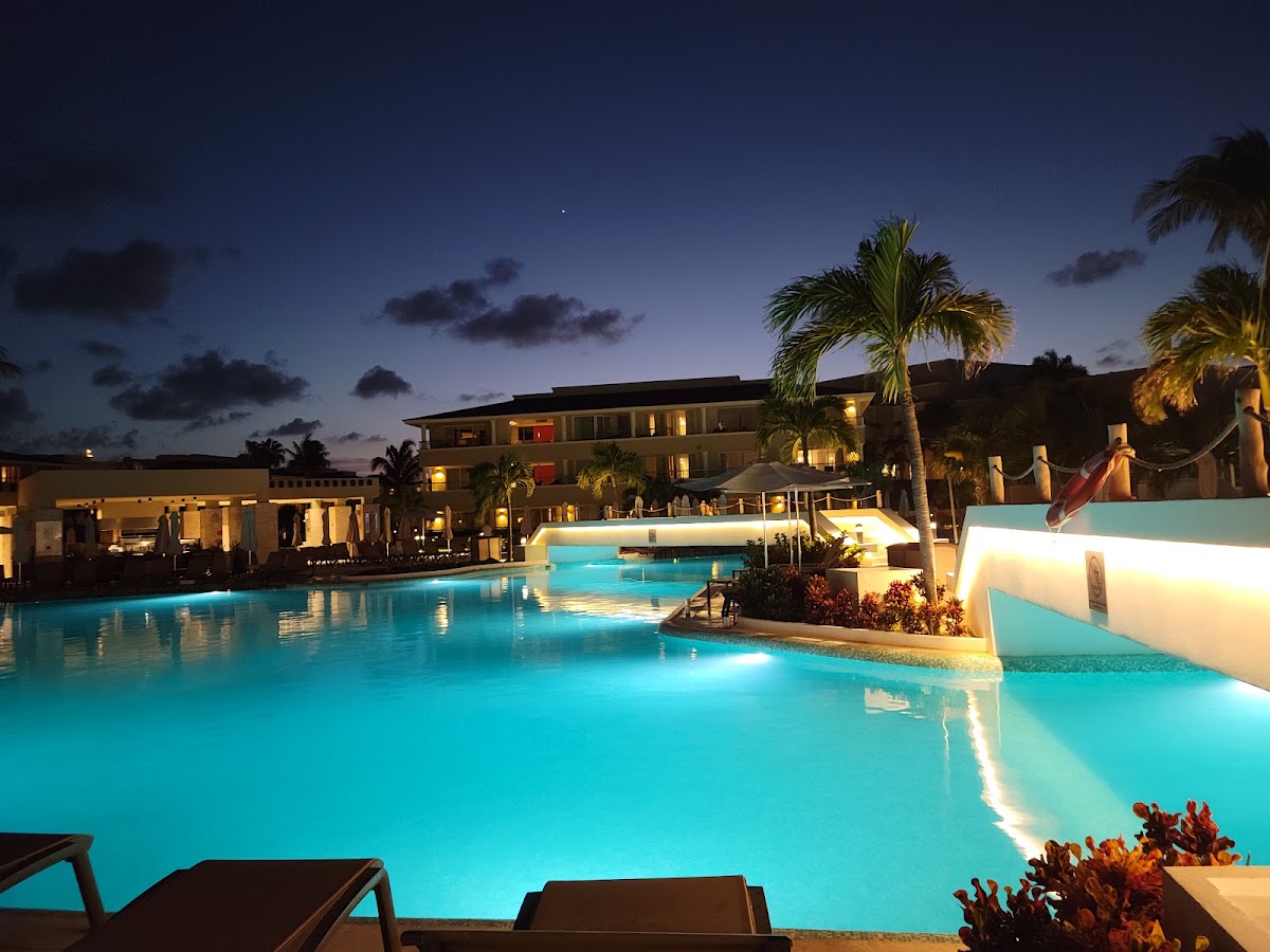 Illuminated turquoise pool complex at dusk with resort buildings, palm trees, and lounge chairs in the foreground at Moon Palace Cancun.