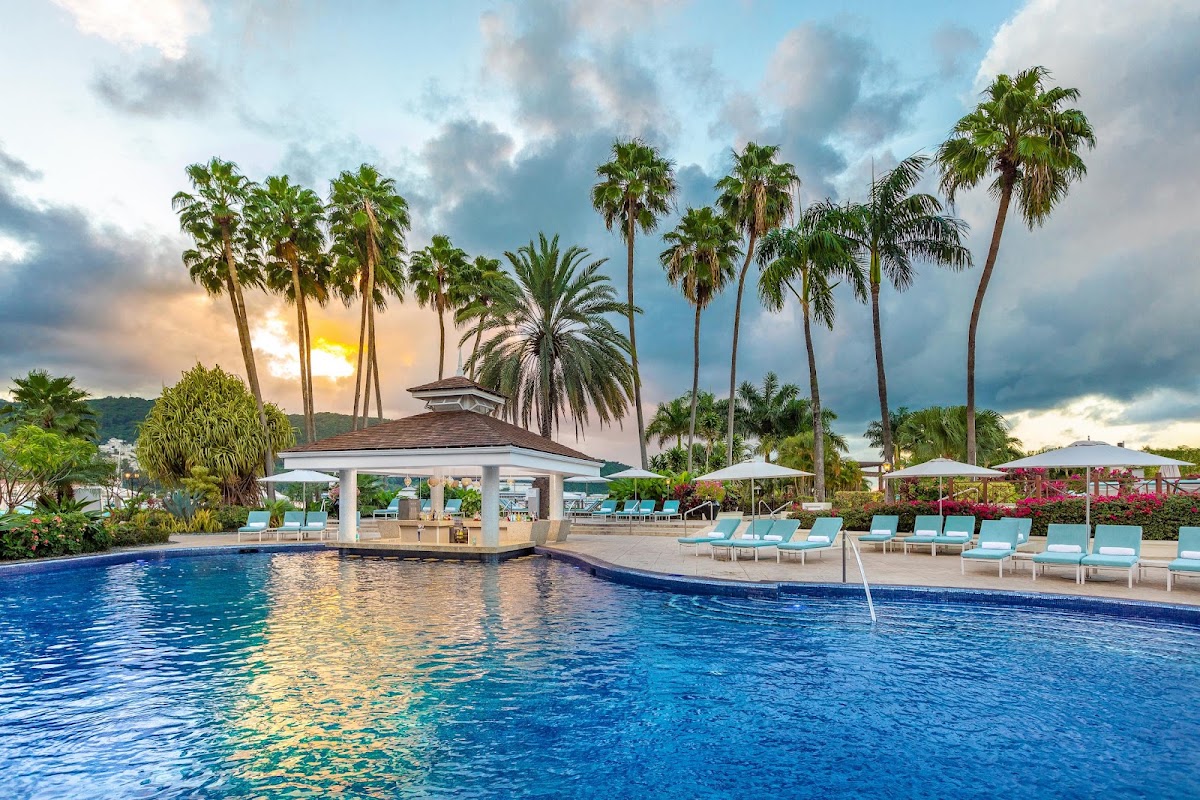 Resort pool with swim-up bar pavilion, lounge chairs, umbrellas, and palm trees at sunset under cloudy sky.