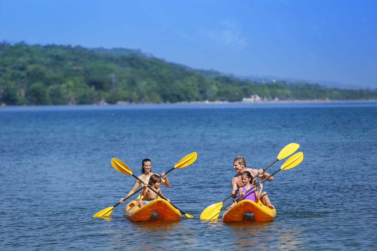 Two families paddle yellow kayaks in calm blue water with forested cliffs and beach in the background.