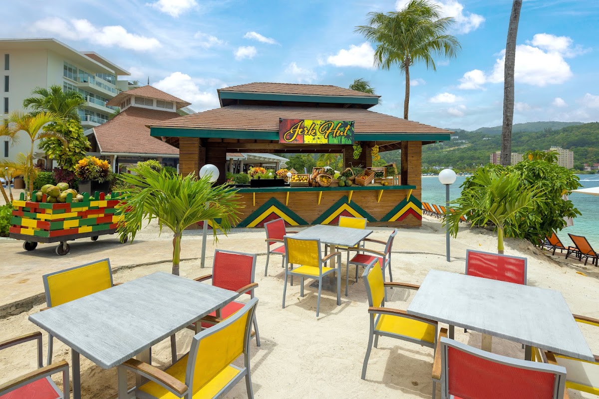 Beach bar with colorful chairs and dining tables at Moon Palace Jamaica, overlooking the ocean and resort buildings.