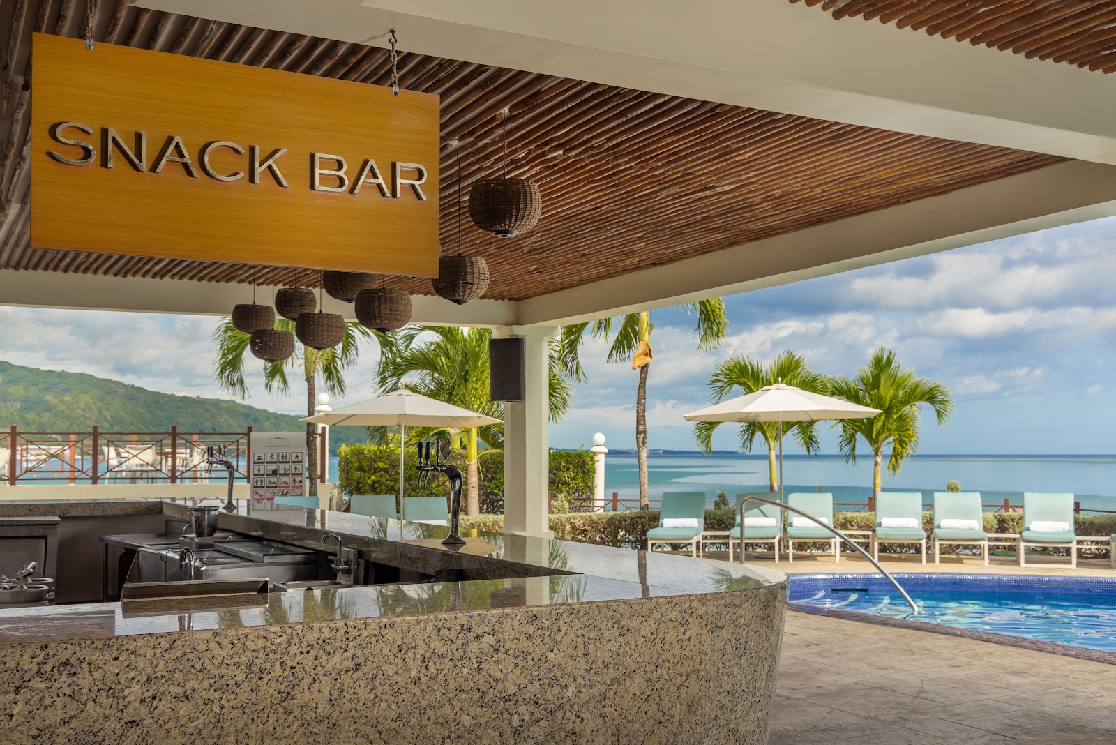 Snack bar counter with ocean view overlooking pool, lounge chairs, and Caribbean Sea beyond palm trees.