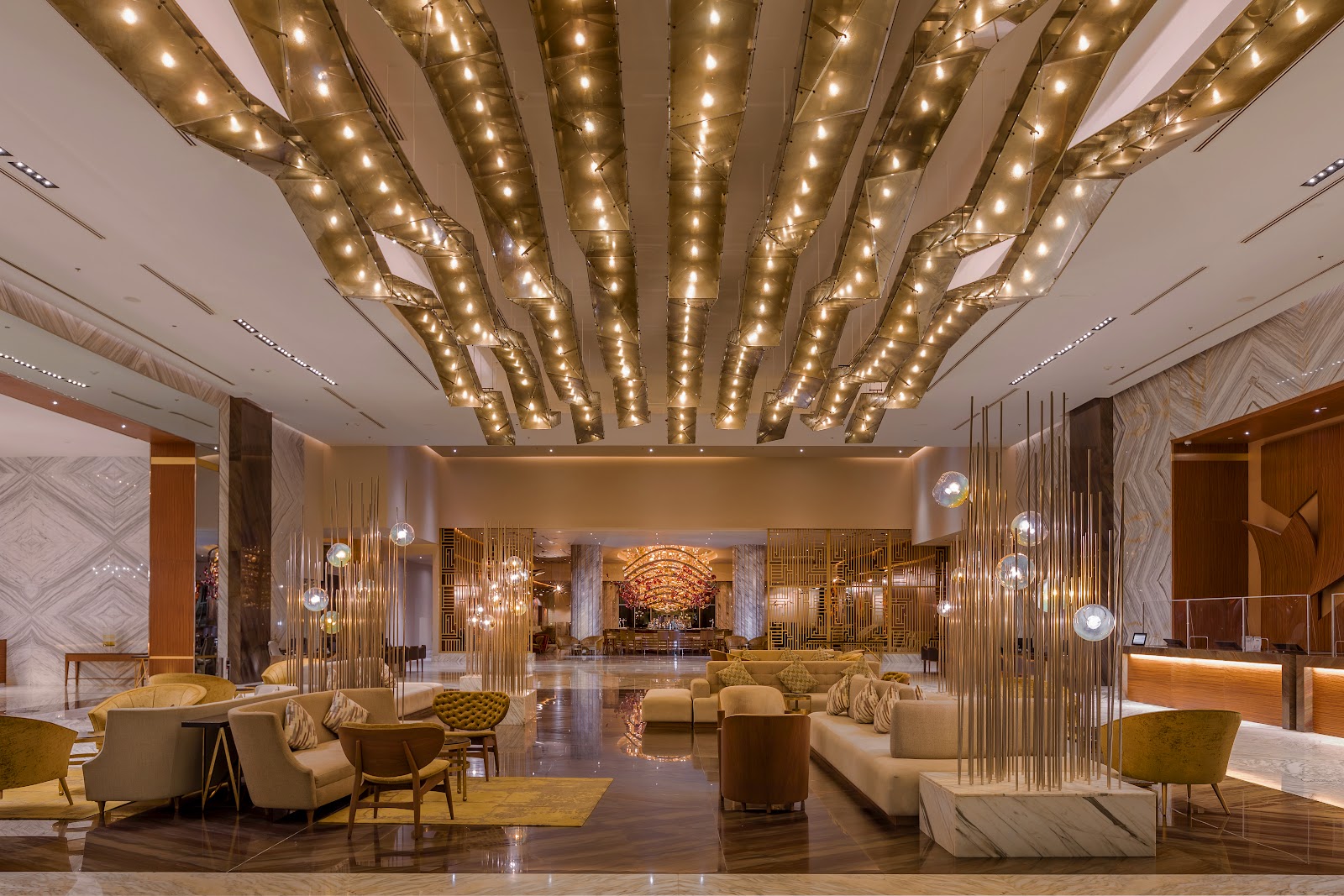 Modern lobby with brass tube lighting fixtures overhead, cream and gold seating areas, marble columns, and polished floors.
