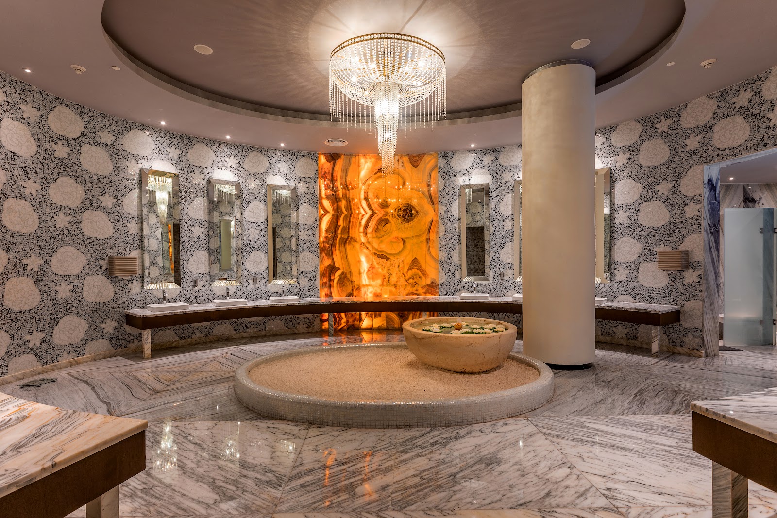 Spa bathhouse with marble floors, patterned walls, crystal chandelier, and glowing orange artwork above a sunken soaking tub with flower petals.