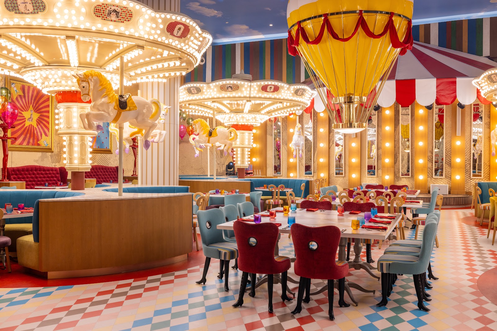 Colorful carnival-themed dining room with illuminated carousel horses, red and gold hot air balloon, striped walls, and tables set for dining at Moon Palace The Grand.