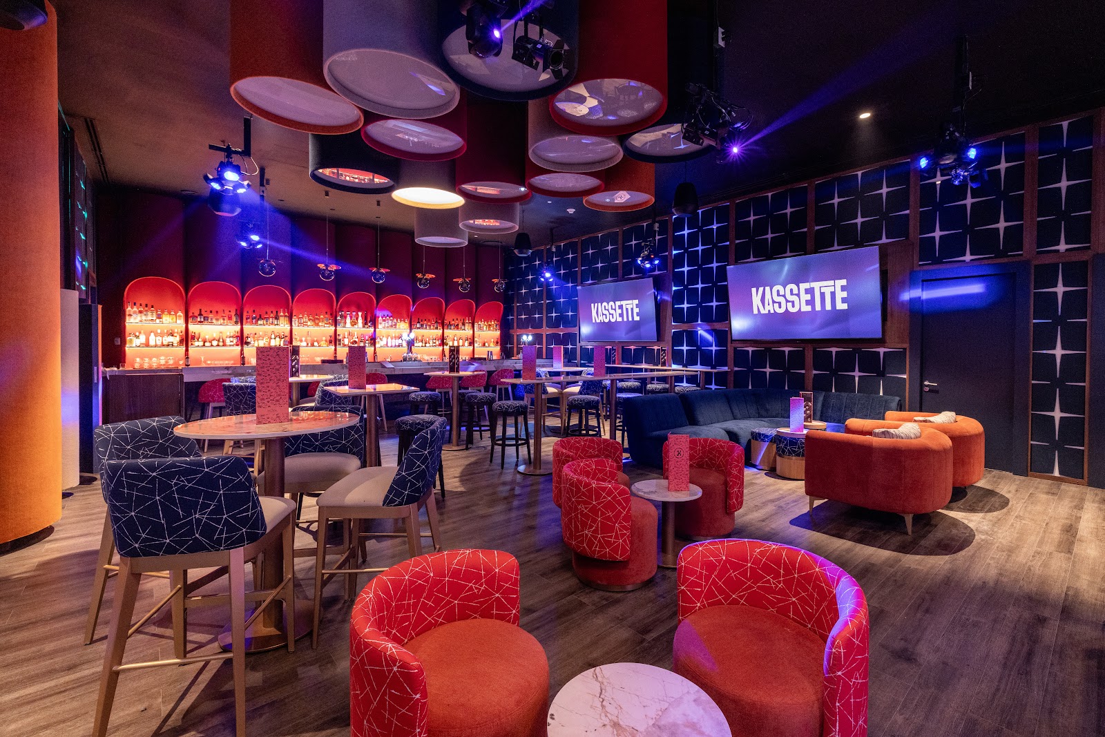 Modern nightclub interior with red and black patterned seating, illuminated ceiling discs, blue accent lighting, and Kassette signage on screens.