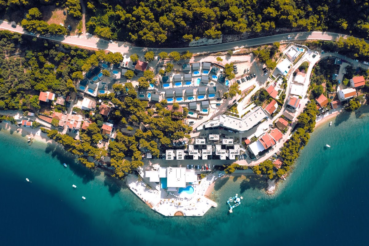 Aerial view of Morenia Beach Resort on turquoise Adriatic water, surrounded by dense forest with terracotta-roofed buildings and blue pools.