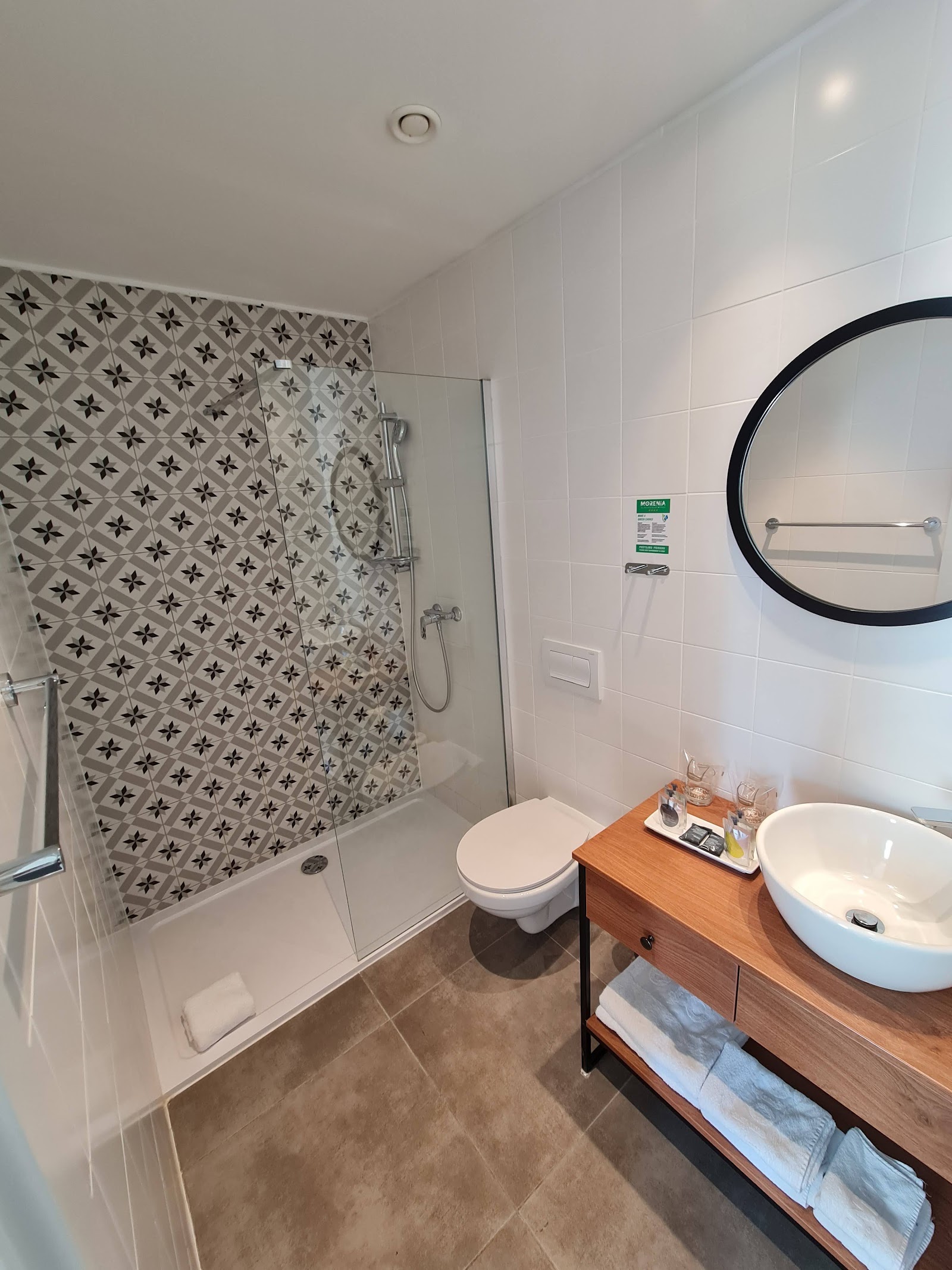 Modern bathroom with patterned shower wall, round mirror, wooden vanity, and white round sink at Morenia Beach Resort.