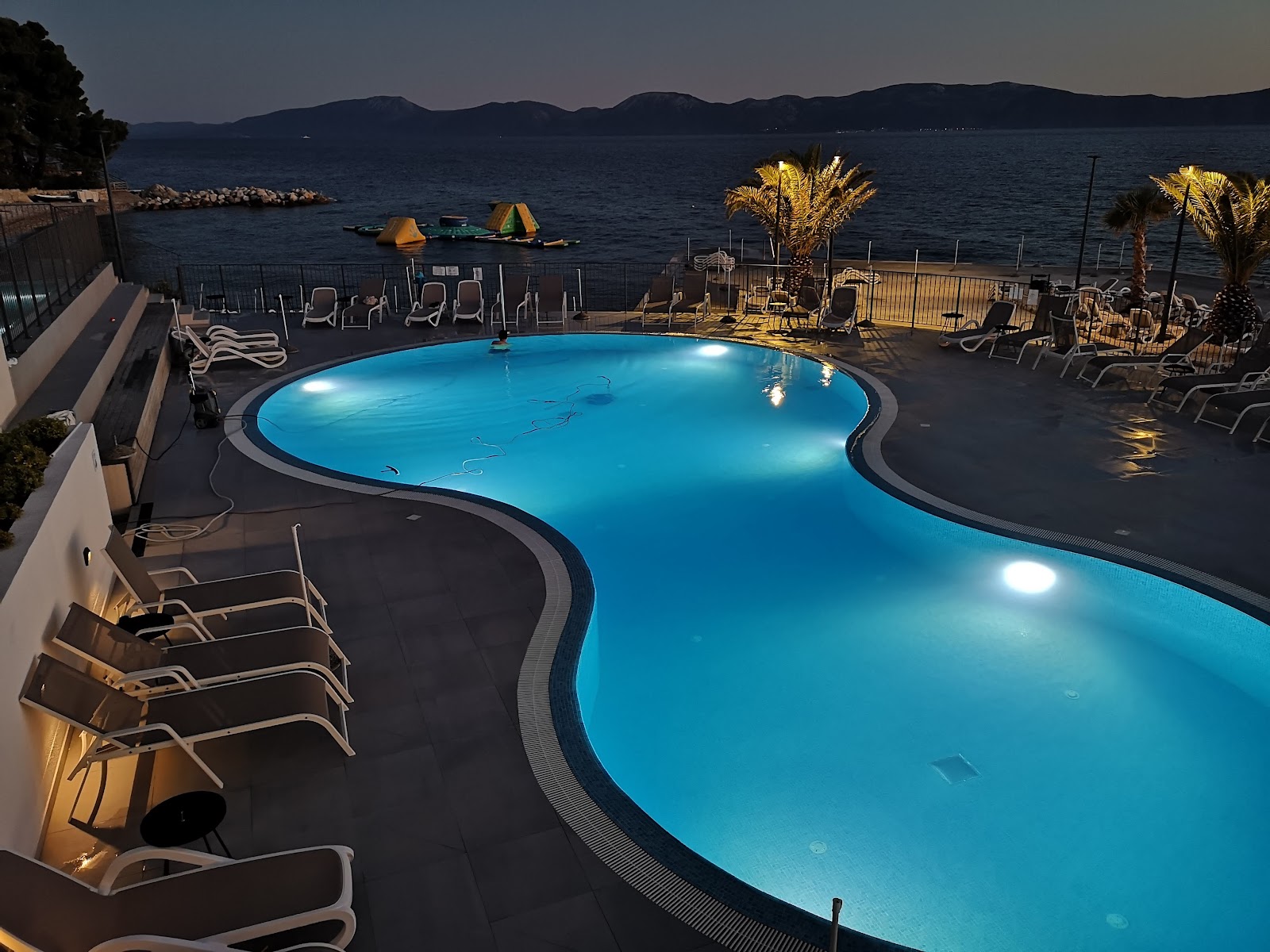 Illuminated pool overlooks the Adriatic Sea at dusk, with loungers, palm trees, and mountains visible across the water.