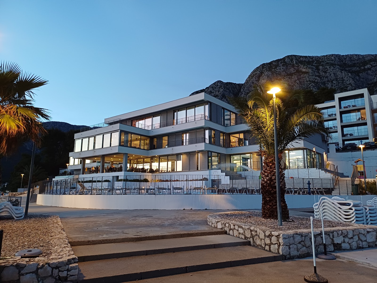 White modern resort buildings nestle on a rocky coastline with turquoise water, pine forest, and dramatic limestone mountains rising behind.