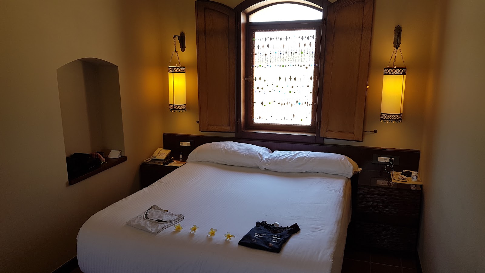 Bedroom with queen bed, white linens, dark wood furniture, yellow walls, hanging lantern sconces, and a shuttered window with ocean view.