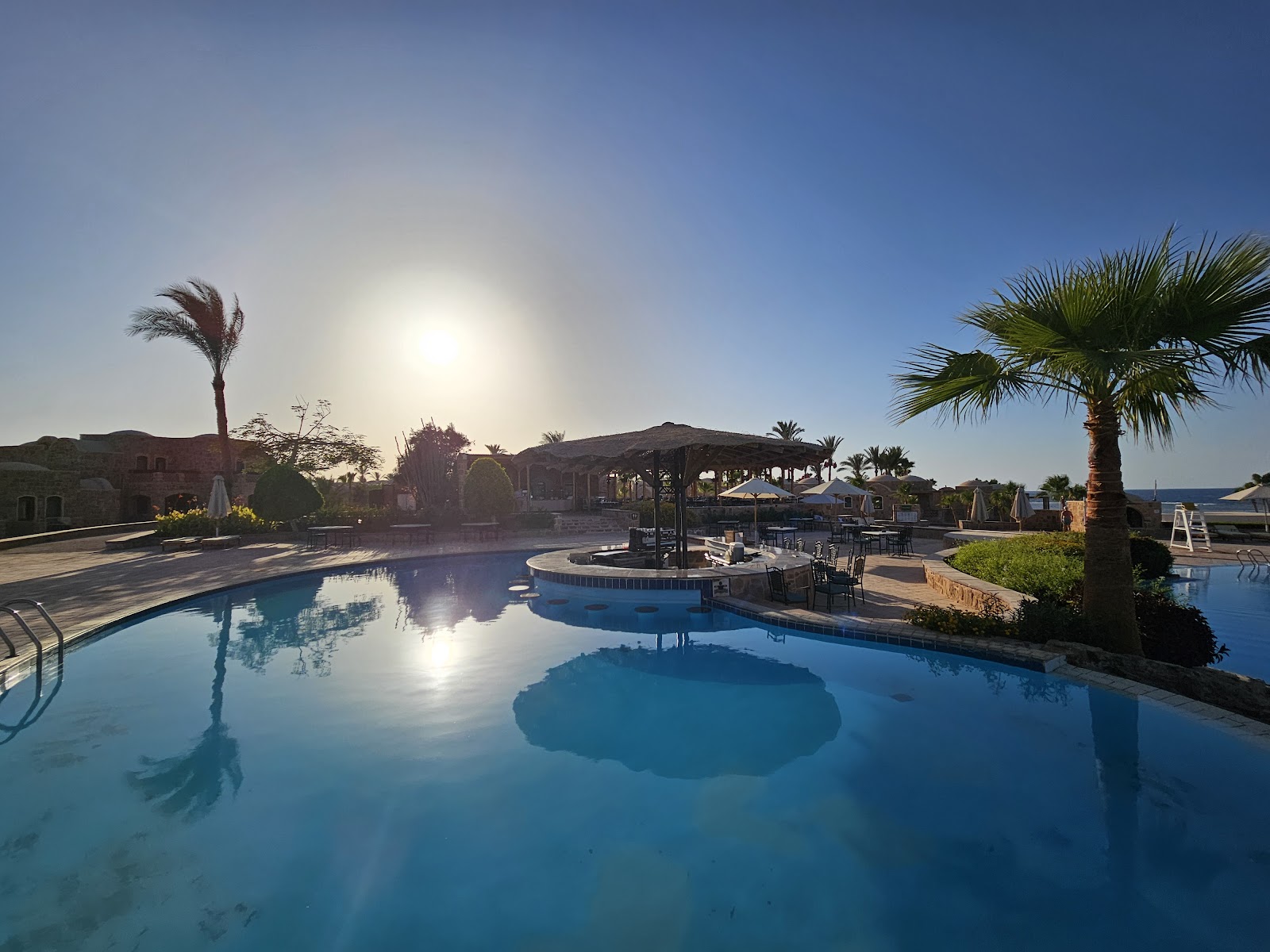 Resort pool at sunrise with central island bar, palm trees, and sea visible in the distance under clear sky.