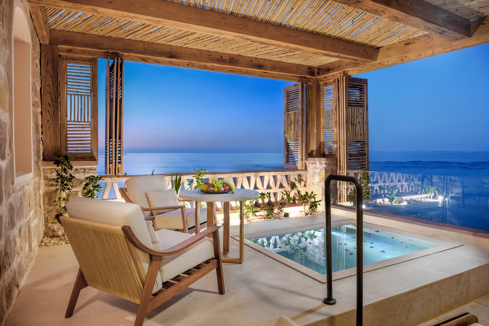 Wooden pergola-shaded terrace with cream armchairs, fruit bowl, and private plunge pool overlooking the calm Red Sea at dusk.