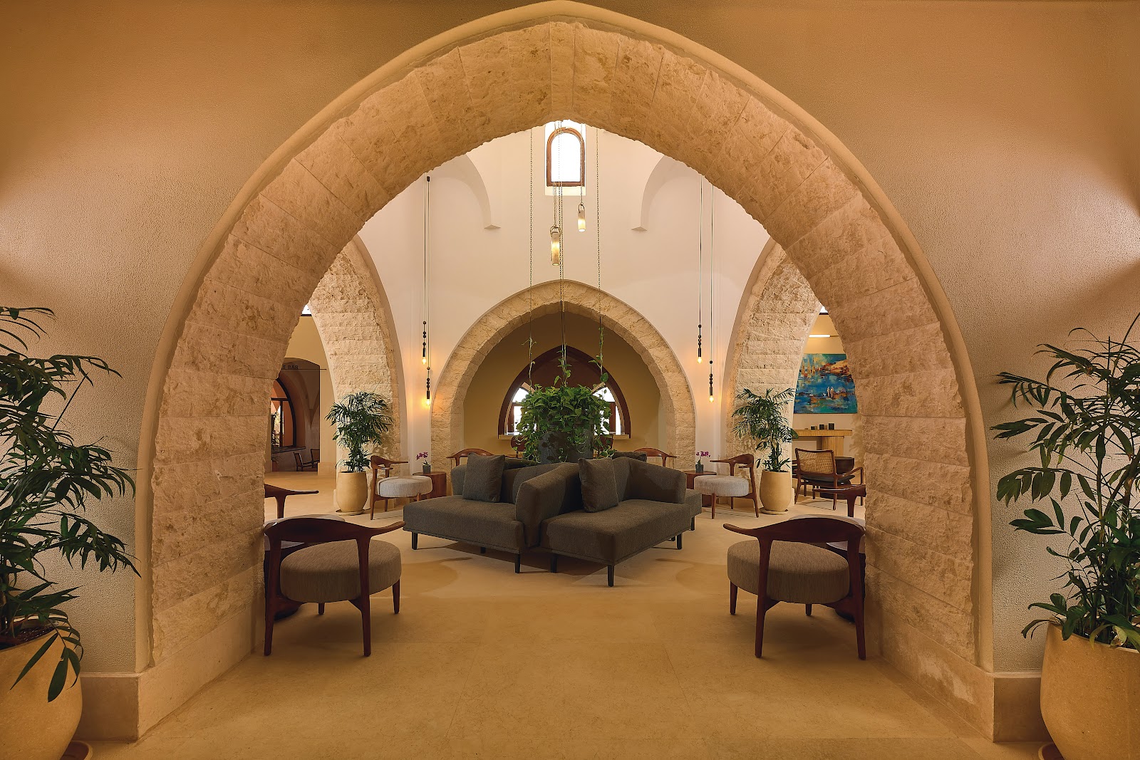 Stone archways frame a lobby lounge with tan seating, potted plants, and hanging pendant lights in earthy tones.