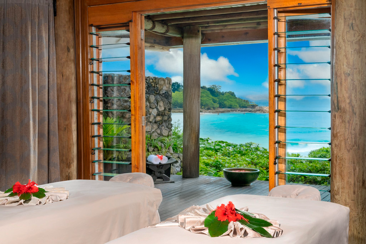Spa treatment room with massage tables decorated with red flowers, opening to a wooden deck overlooking turquoise ocean and green hillside at Namale Resort & Spa.