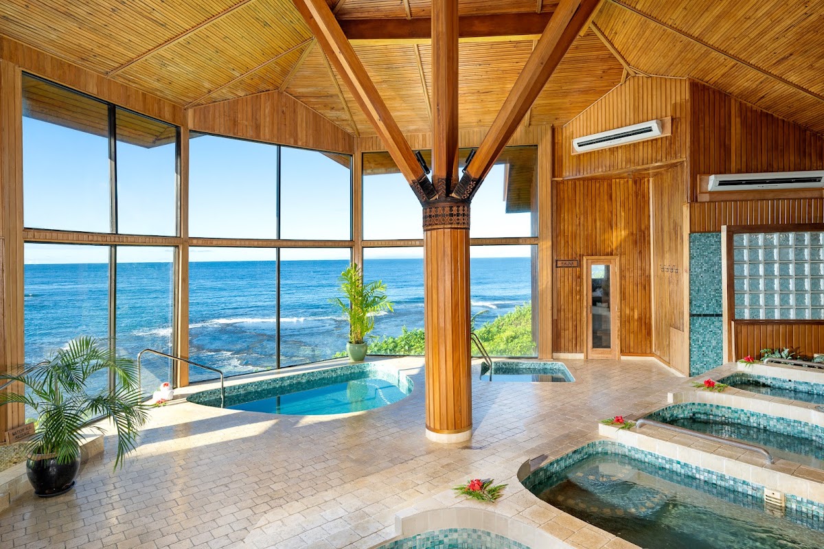 Spa interior with wooden beams and glass walls overlooking the ocean, featuring multiple tiled soaking tubs and a central pool.