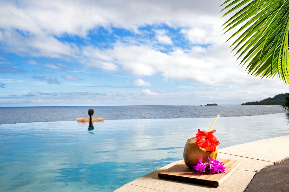 Coconut drink with red hibiscus flower on pool deck at Namale Resort & Spa, with swimmer in infinity pool overlooking Fiji ocean and islands.