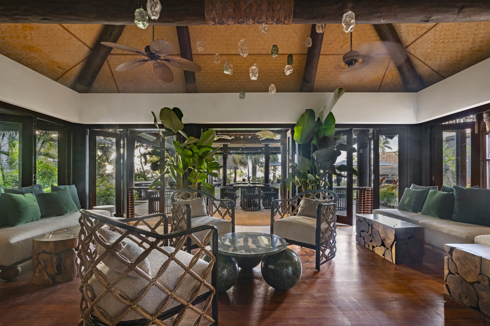 Indoor lounge at Nanuku Resort Fiji with wooden ceiling, hanging glass accents, tropical plants, and open views to the surrounding garden and ocean beyond.