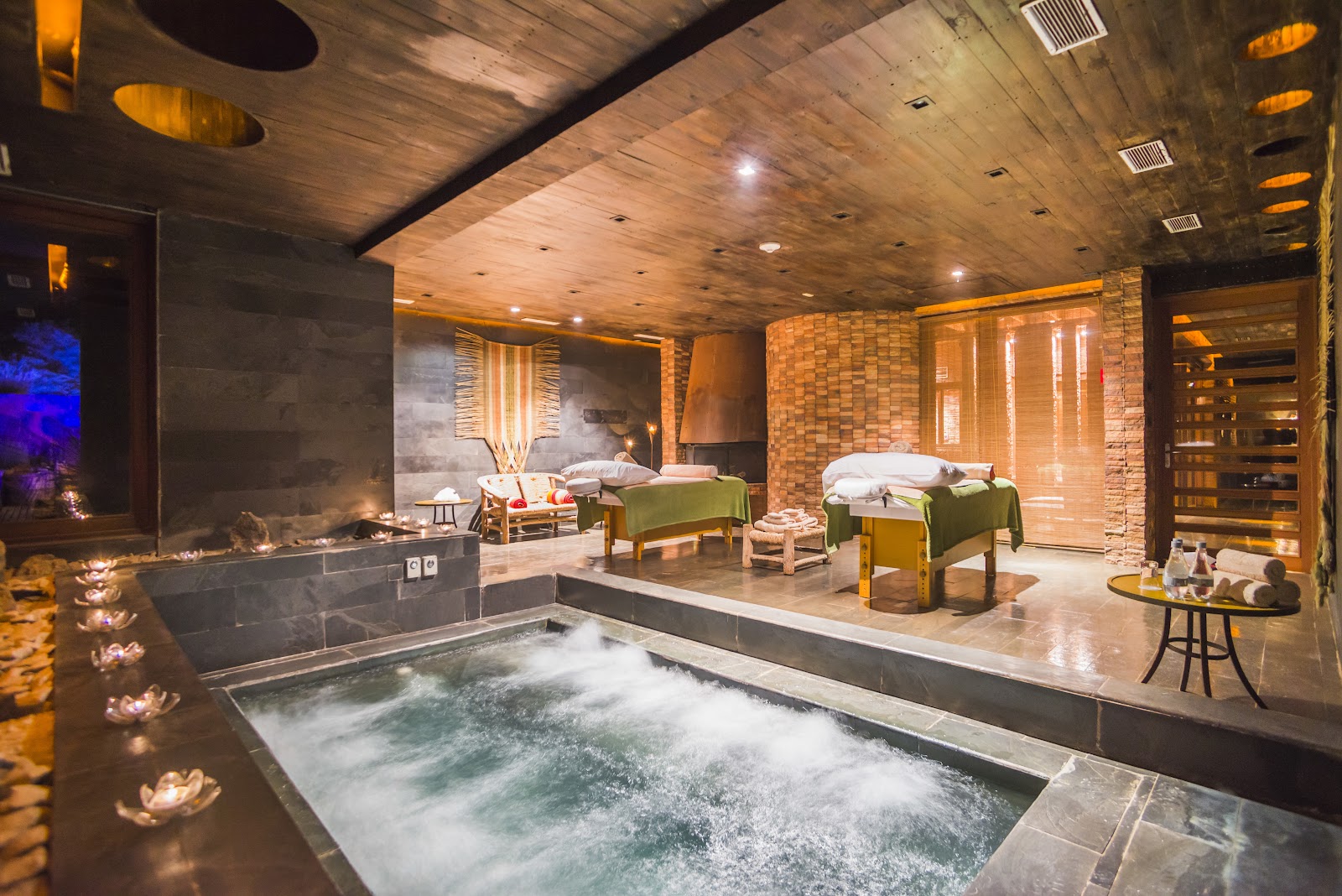 Indoor spa with bubbling thermal pool, massage tables with green cushions, brick walls, wooden ceiling, and fireplace with candles.