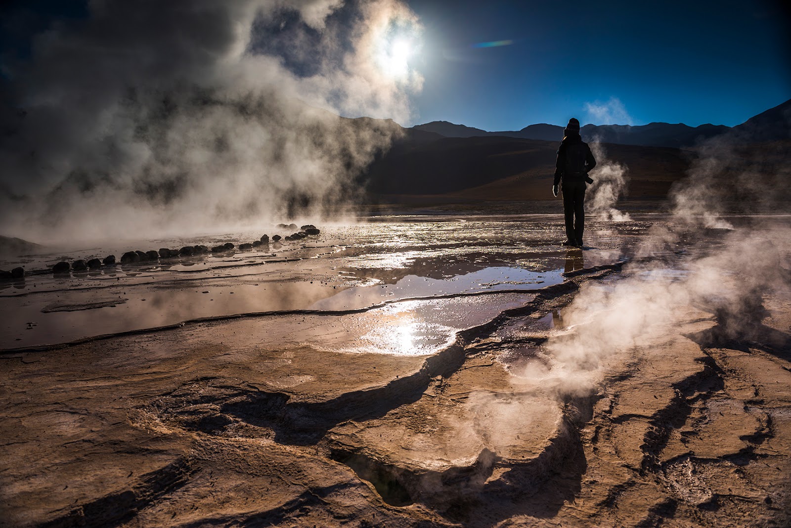 Guest stands among steaming geothermal pools and hot springs in the Atacama Desert with mountains in the background.