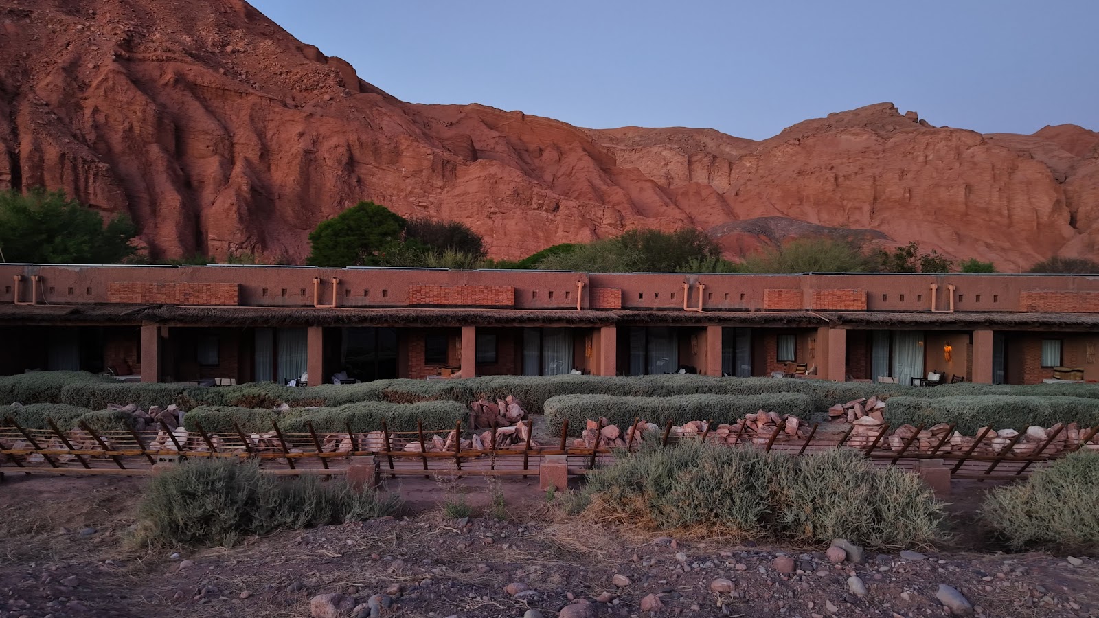 Terracotta-colored bungalows with shuttered windows line a desert courtyard dotted with lounge chairs and shrubs beneath red rock mountains.
