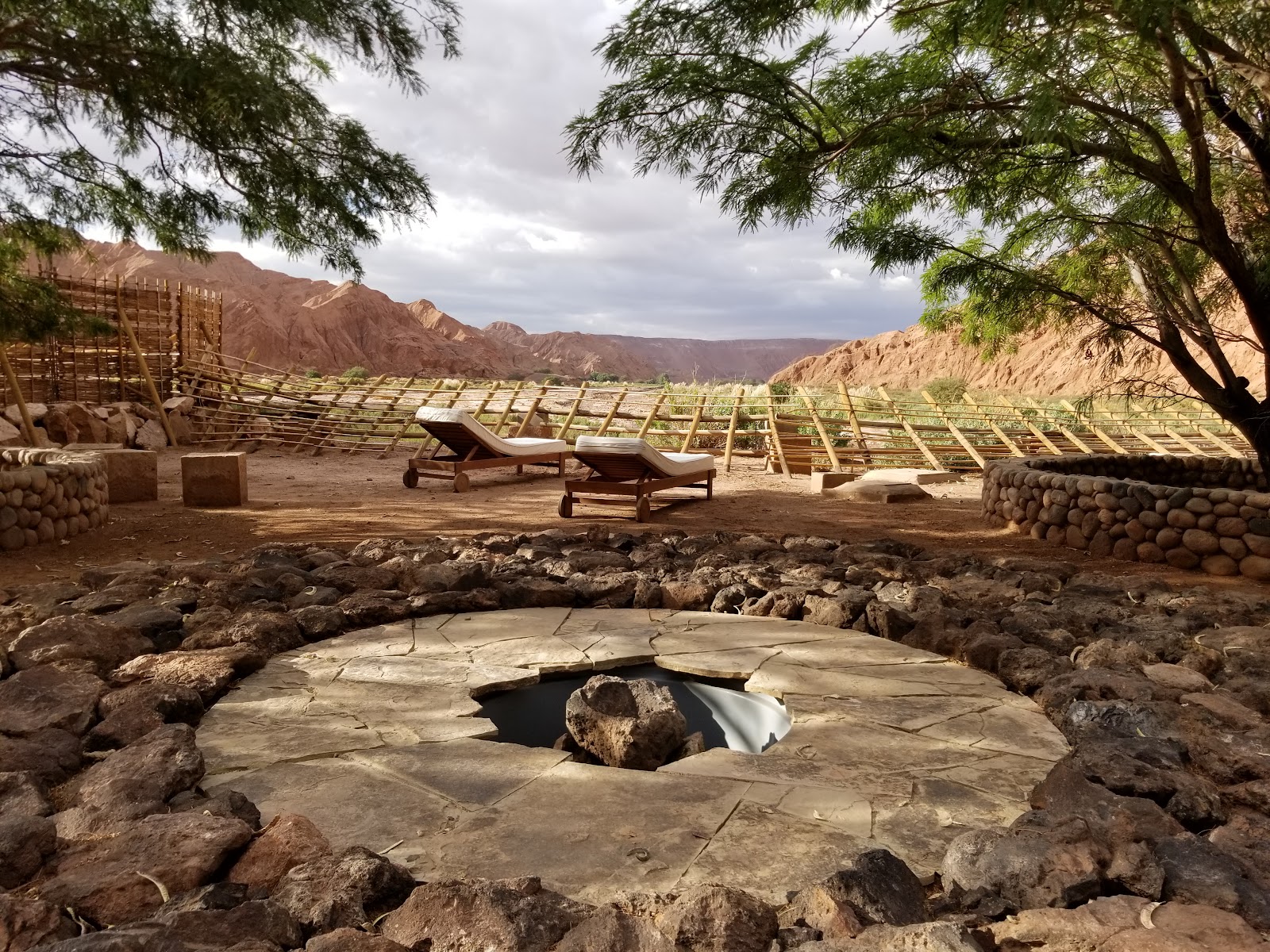 Hot spring pool with stone surround in desert valley, lounge chairs and acacia trees visible under rugged mountains at Nayara Alto Atacama.