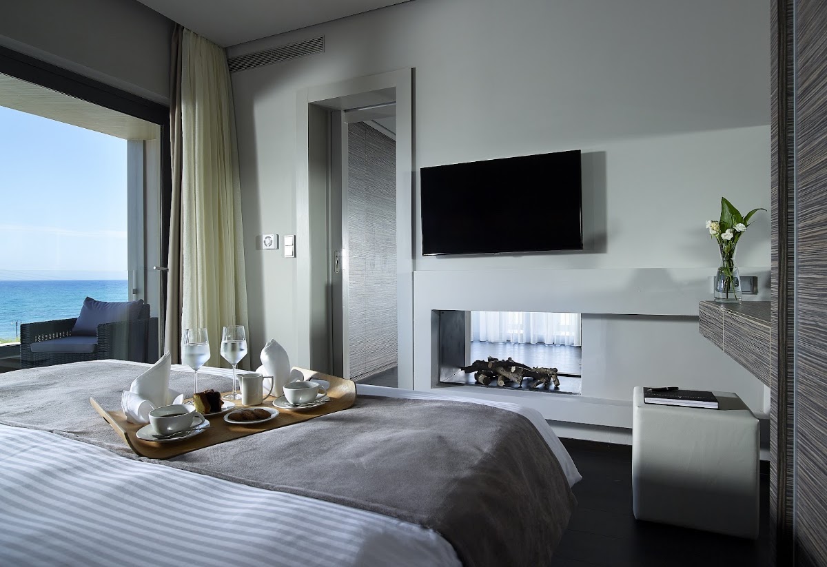 Modern resort bedroom with wall-mounted TV, electric fireplace, and ocean view through floor-to-ceiling windows with balcony seating.