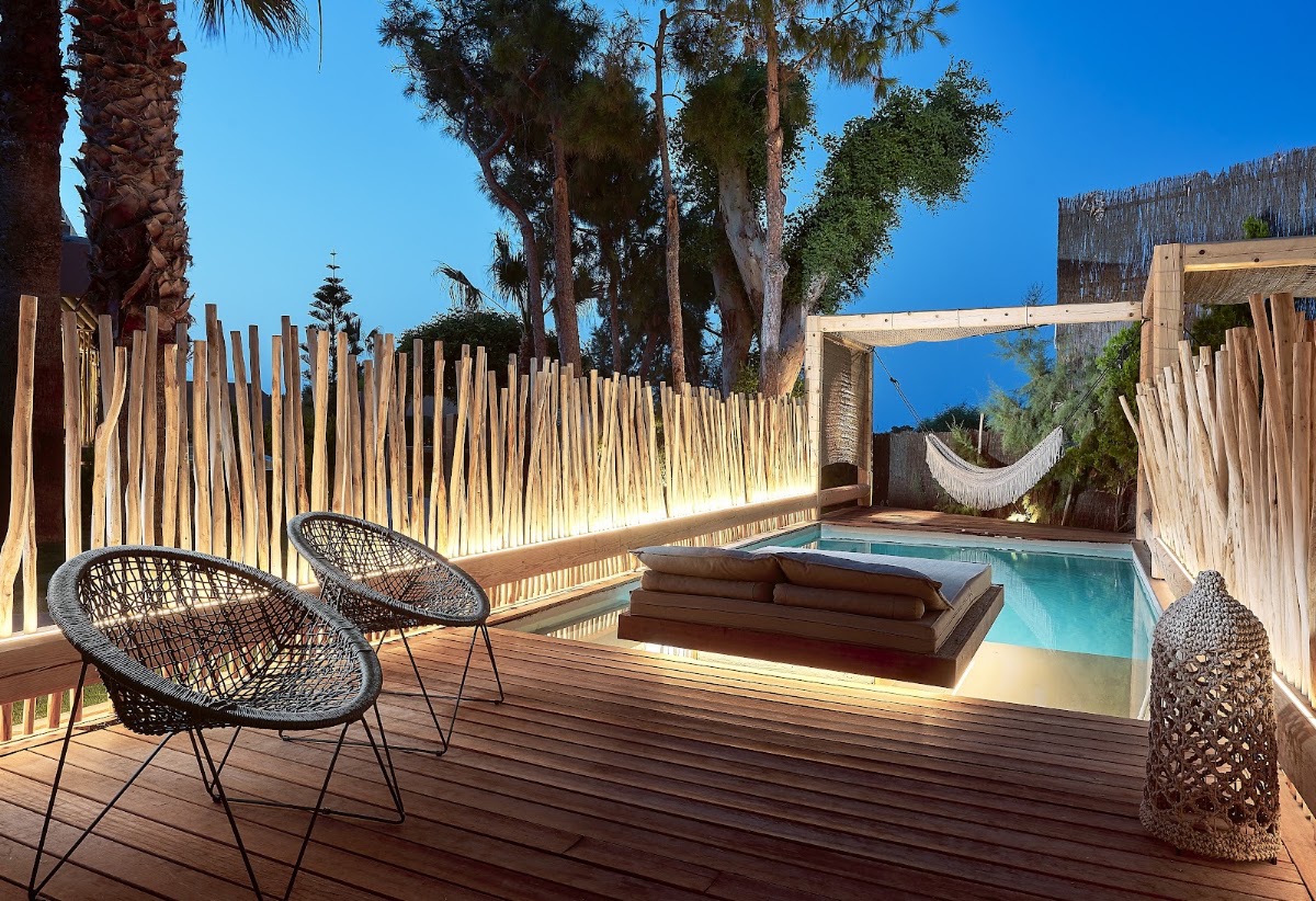 Private villa terrace with woven lounge chairs, small plunge pool, wooden deck, palm trees, and woven privacy screens under clear blue sky.