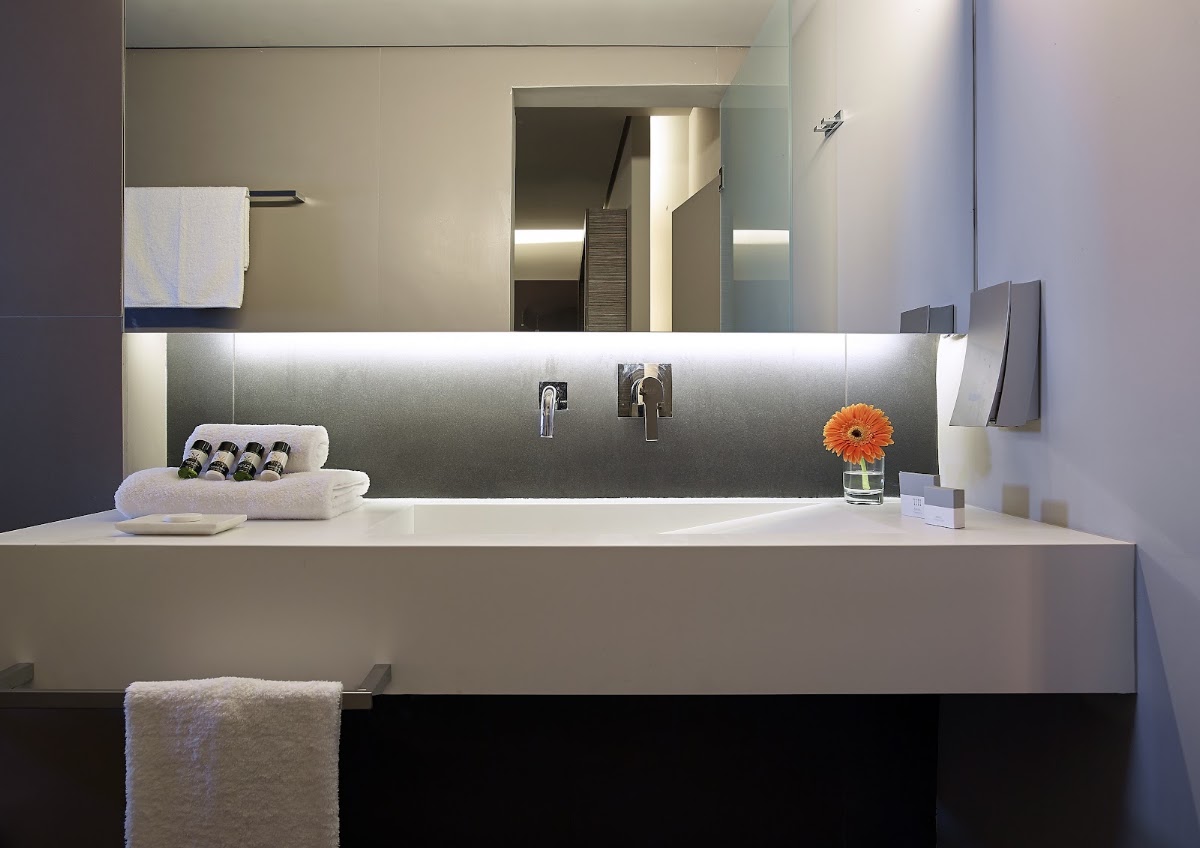 Modern bathroom vanity with white countertop, dual chrome faucets, stacked white towels, orange flower in glass, and illuminated mirror above.