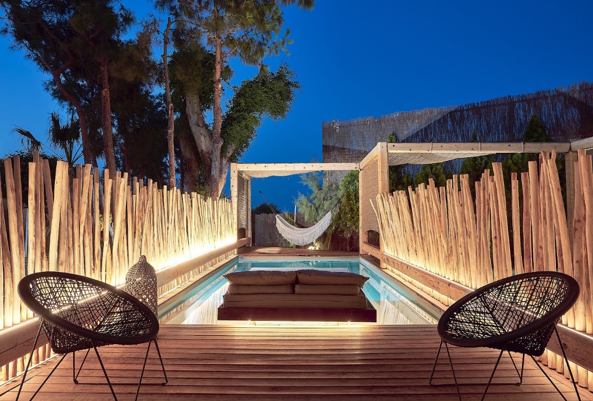 Wooden deck with two woven black chairs facing a narrow pool lined with loungers, bordered by tall wooden screens and palm trees under clear blue sky.