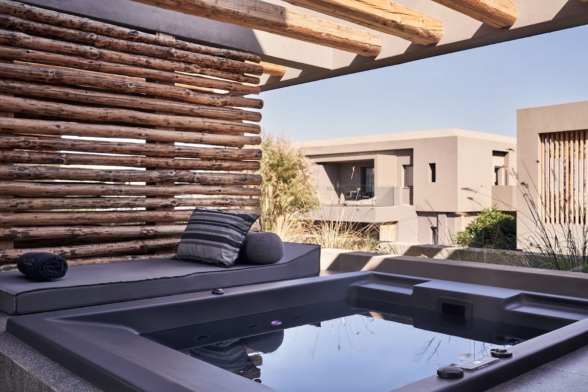 Sunlit courtyard with sunken hot tub, wooden pergola, dark lounger with pillows, and modern beige buildings at NEMA Design Hotel & Spa.
