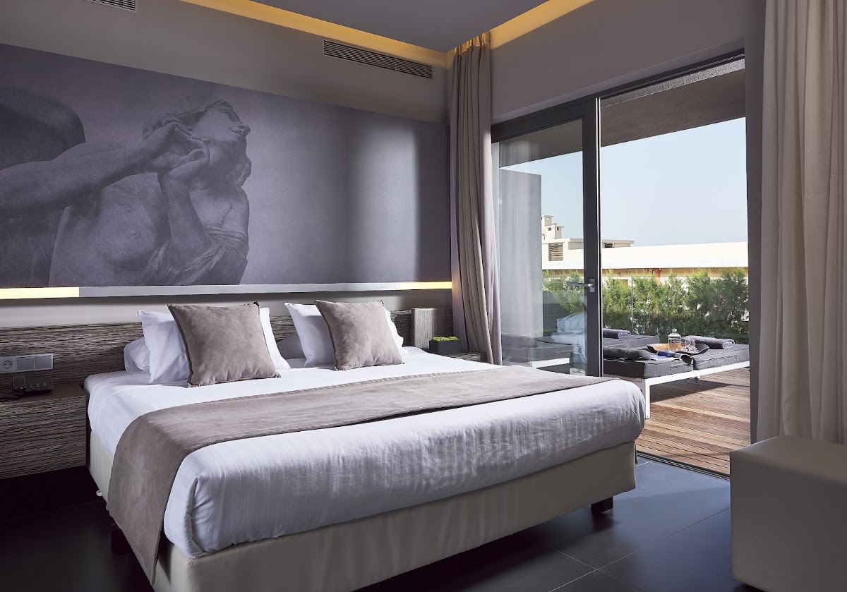 Modern hotel bedroom with king bed, sculptural wall art, and sliding glass doors opening to a terrace with loungers and sea view.